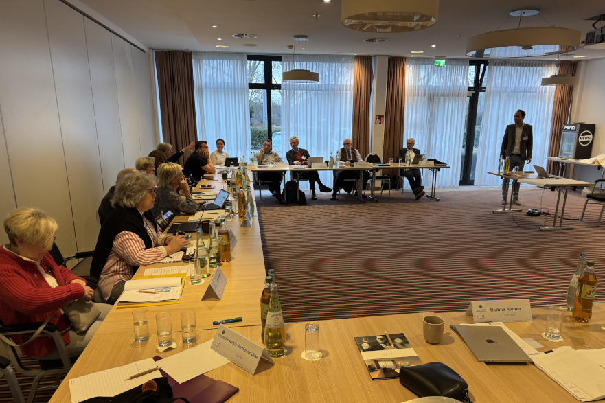 Foto eines Seminarraums mit Teppich ausgelegt. Tische stehen in U Formation, mehere Teilnehmer, Tische sind gefüllt mit Blocken und Stiften, die Teilnehmer lauschen Laurent Wagner, dem Programmreferenten, der einen Vortrag hält.